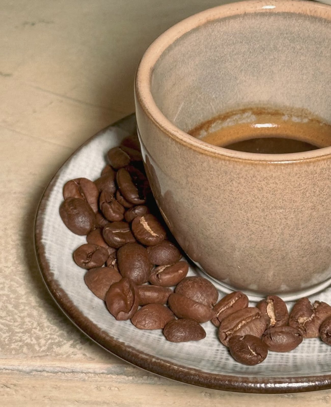 An espresso in a stoneware cup on its saucer, surrounded by a scatter of roasted coffee beans