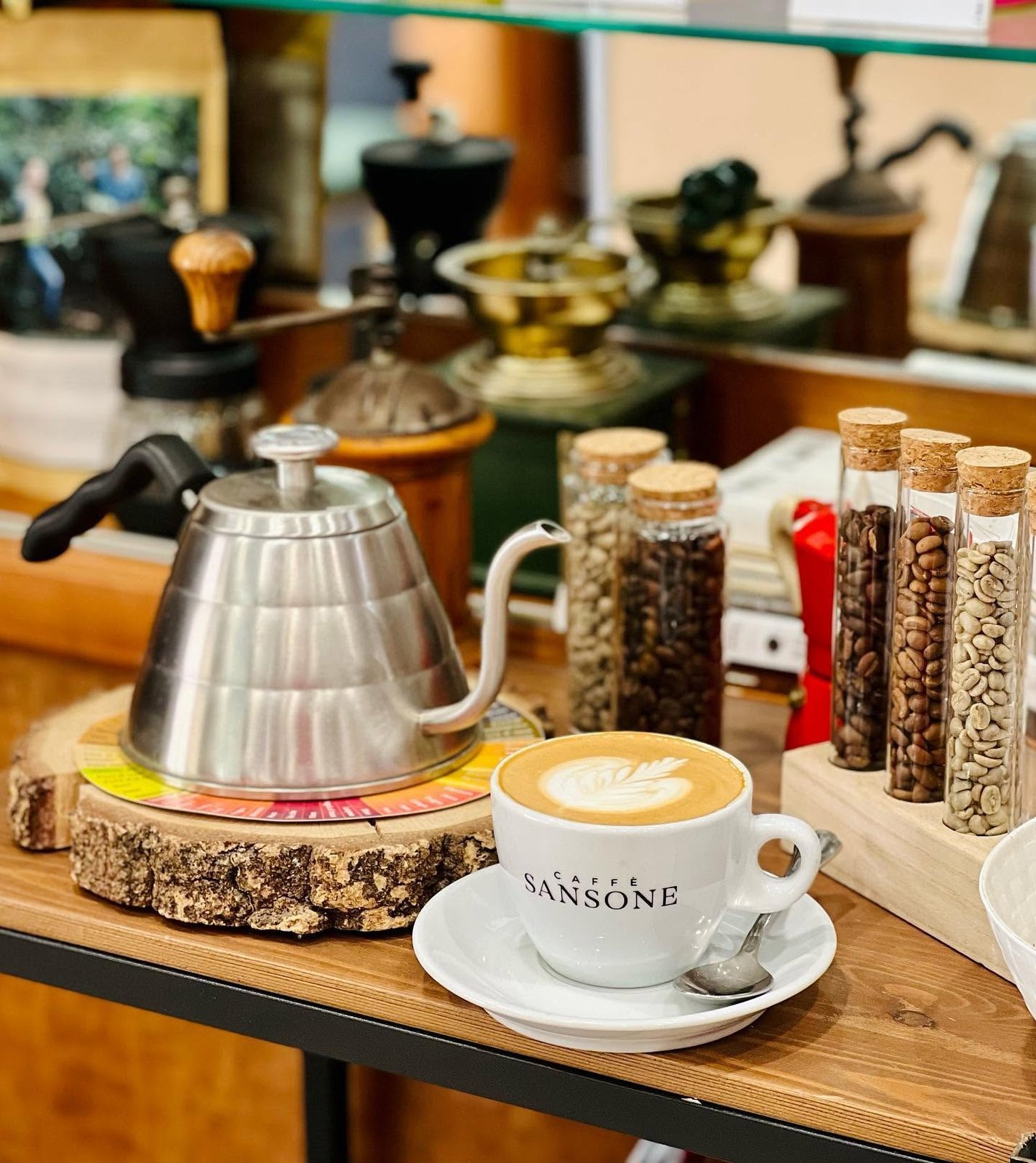 A latte with leaf shaped art in a Caffè Sansone branded cup on the roastery counter, next to a pour over kettle and glass tubes of coffee beans