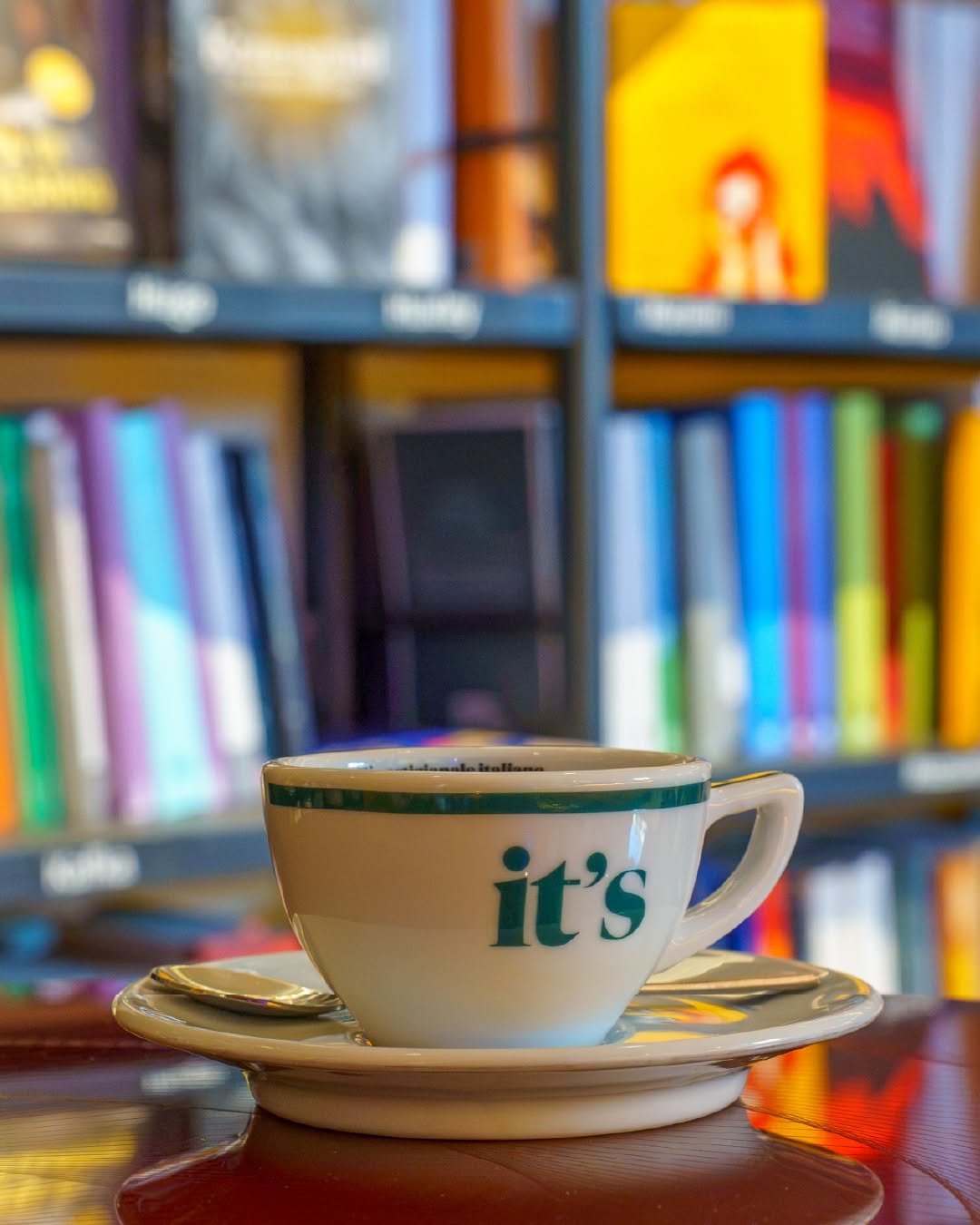 A cup and saucer branded with the It's logo, set on a wooden table against a colorful bookshelf background