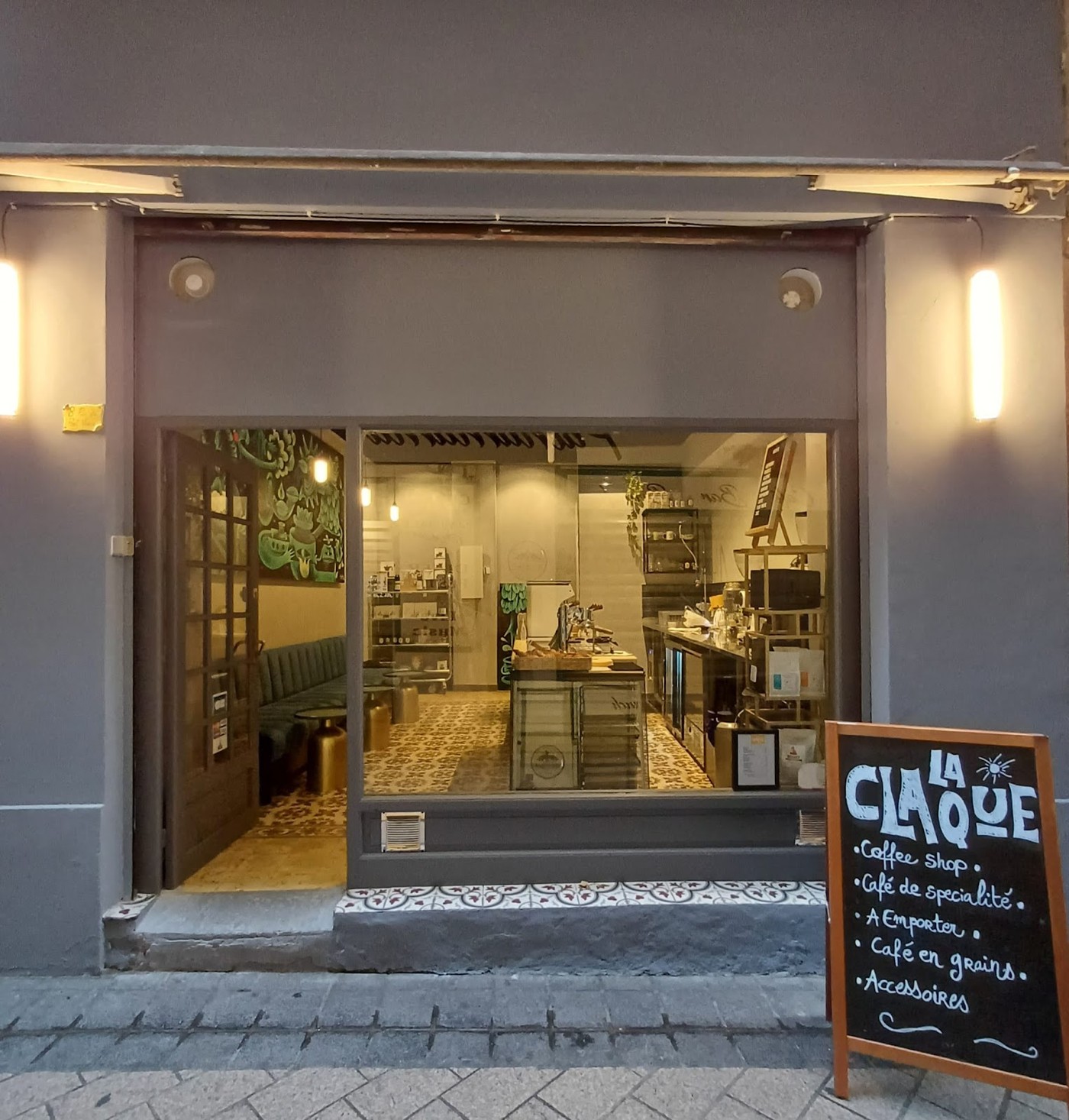 Storefront of a small coffee shop on a narrow old town street, with an illuminated interior visible through the window and a chalkboard sign reading Coffee shop and Café de spécialité out front