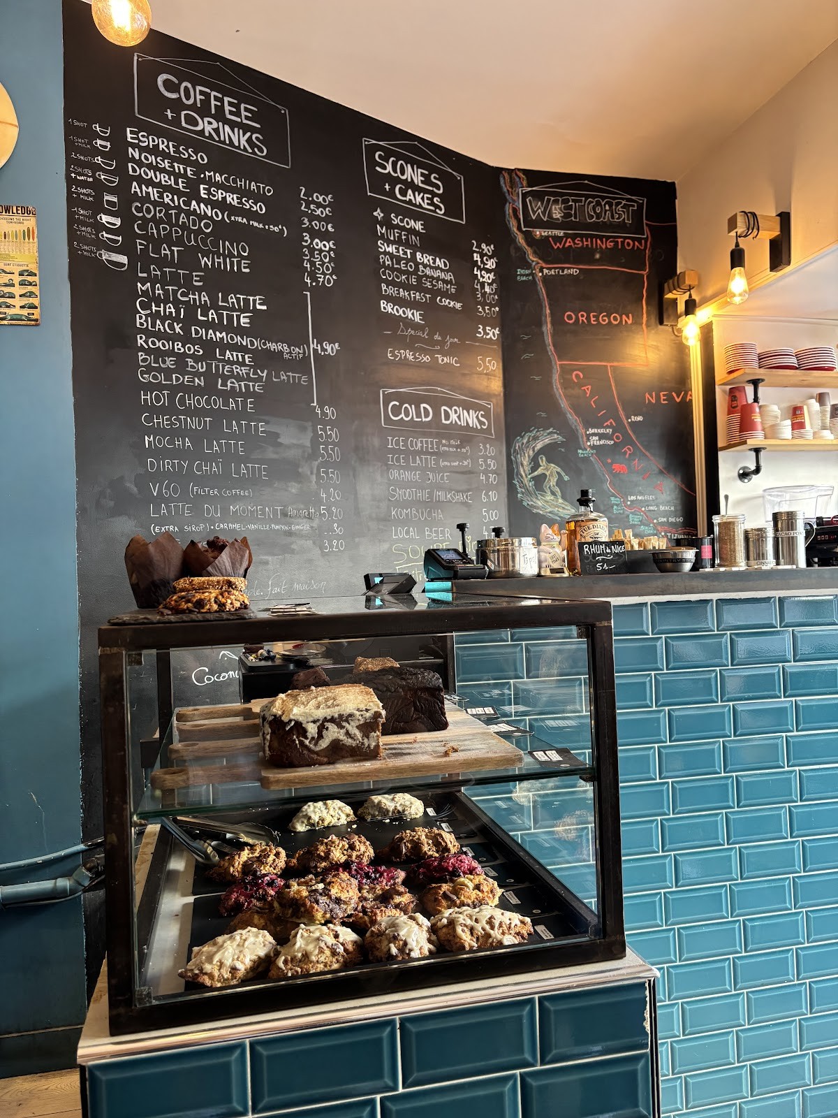Interior counter of a coffee shop with a hand drawn chalkboard menu and West Coast map mural, a pastry display case with scones and banana bread in front, and blue tiled walls