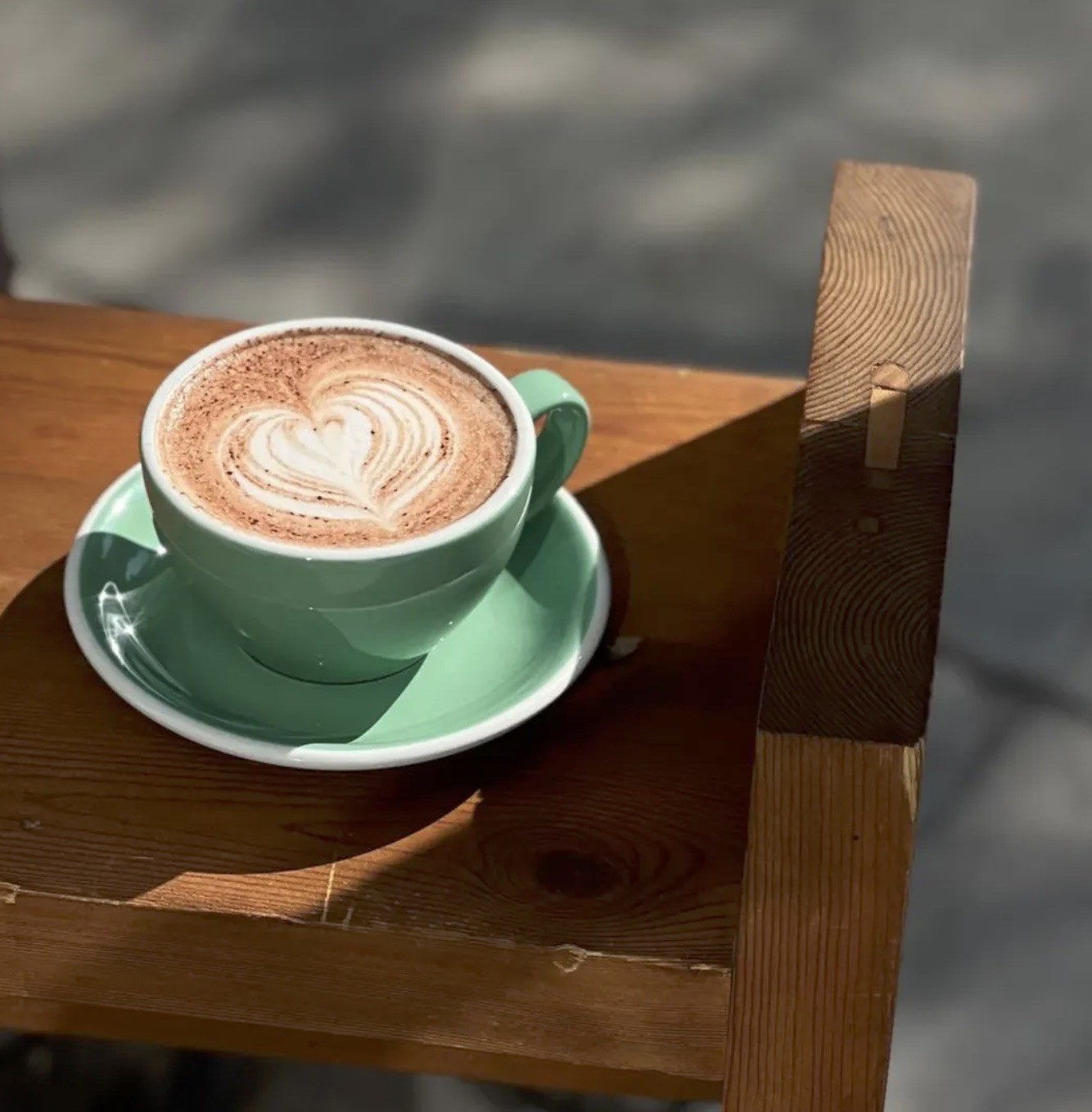 A cappuccino with heart shaped latte art in a mint green cup and saucer, resting on a wooden bench in the sun