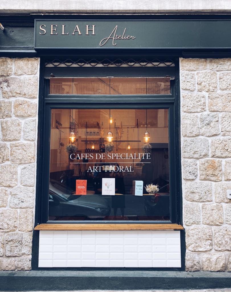 Storefront window of a stone building reading Selah Atelier, Cafés de spécialité, Art floral, with pendant lights and dried flower arrangements visible inside