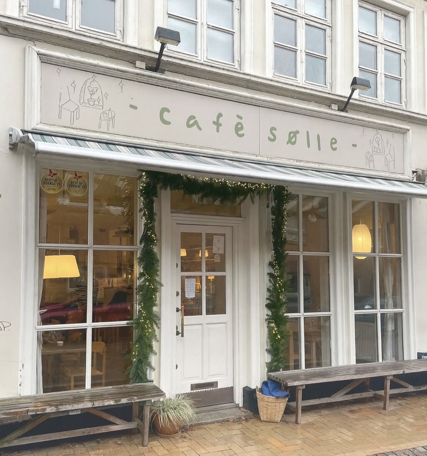 The cream colored storefront of Café Sølle with its hand painted sign, striped awning, and string lit garland framing the door