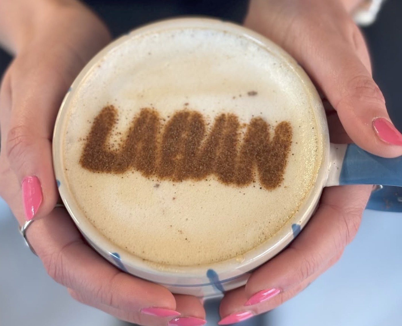 A latte with cinnamon dusted LABAN lettering in the foam, held in two hands