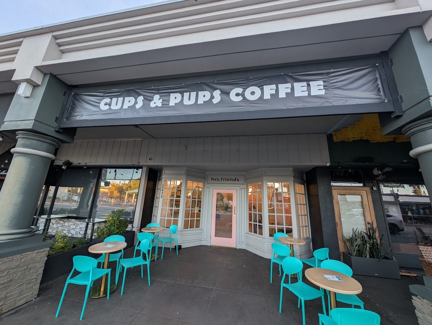 The Cups and Pups Coffee storefront sign above a pink door, with teal outdoor cafe chairs set on the sidewalk patio