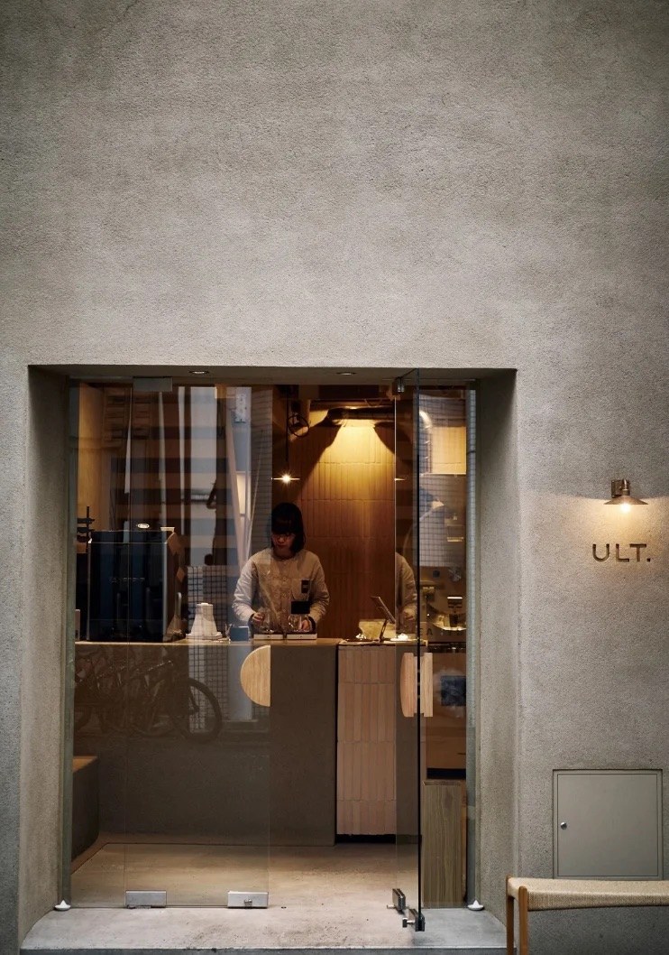 The glass entrance to Ult Coffee with the ULT wall lettering outside and a barista working the counter inside