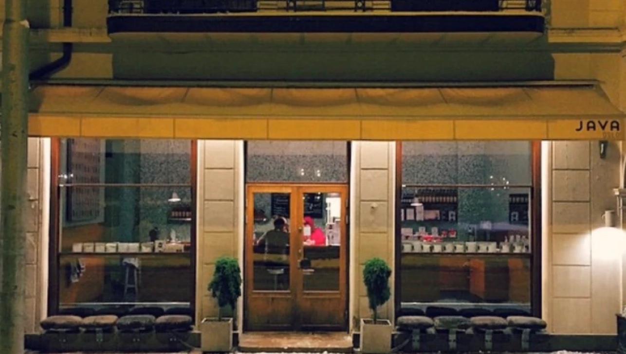 The lit Java Espressobar frontage at night, JAVA lettered on the yellow fascia above tall windows, upholstered stools along the sill and two bay trees flanking the wooden double doors