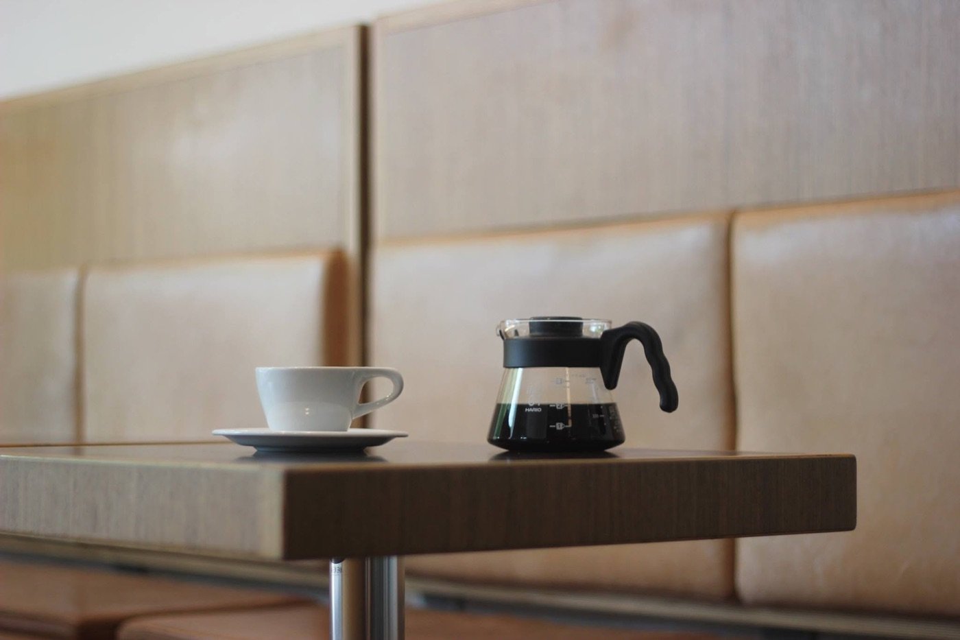 A cup and a Hario carafe of filter coffee on a wooden table against a leather paneled banquette at Mocca
