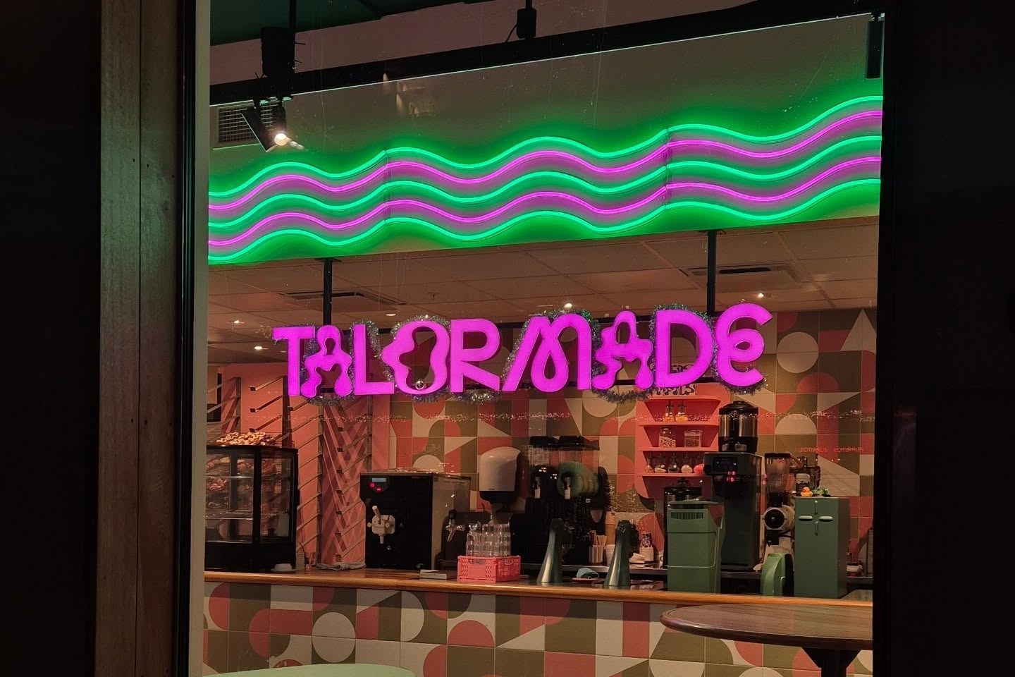 Talormade's pink neon sign glowing above the counter at night, seen through the storefront window with a green neon wave overhead
