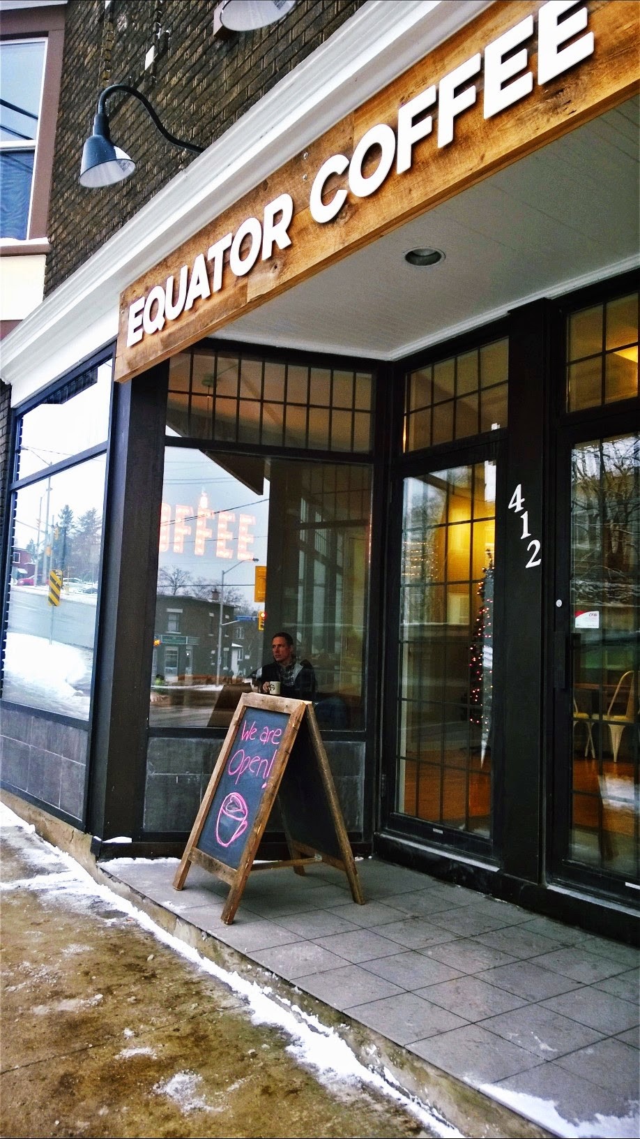 Exterior of Equator Coffee at 412 Churchill Avenue North, with a wood plank EQUATOR COFFEE sign over large glass storefront windows and a sidewalk chalkboard reading We are Open