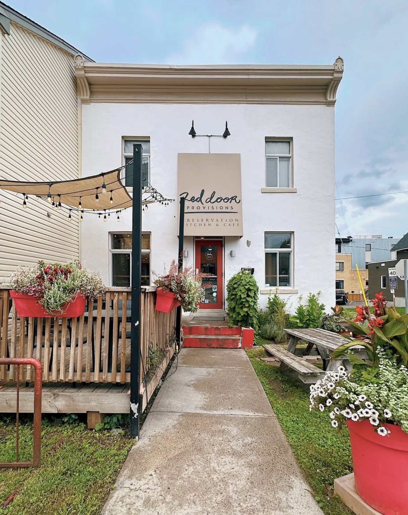 The white stucco exterior of Red Door Provisions with a red front door, red painted steps, string lights over the deck, and flower planters lining the entrance