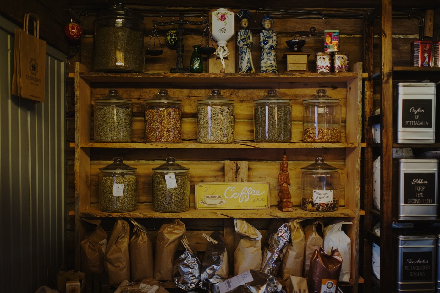 Wooden shelves inside a historic granary shop lined with glass jars of loose tea, a vintage coffee grinder, tea tins, and a hanging Coffee Shop sign