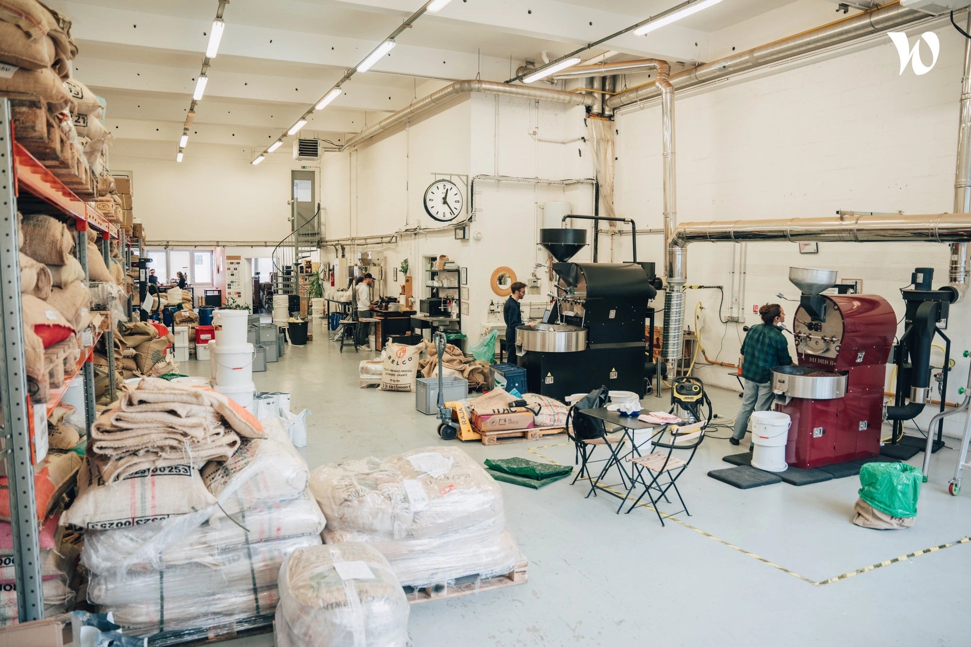 The shared roasting floor at The Beans on Fire, sacks of green coffee stacked beside two roasting machines with roasters at work