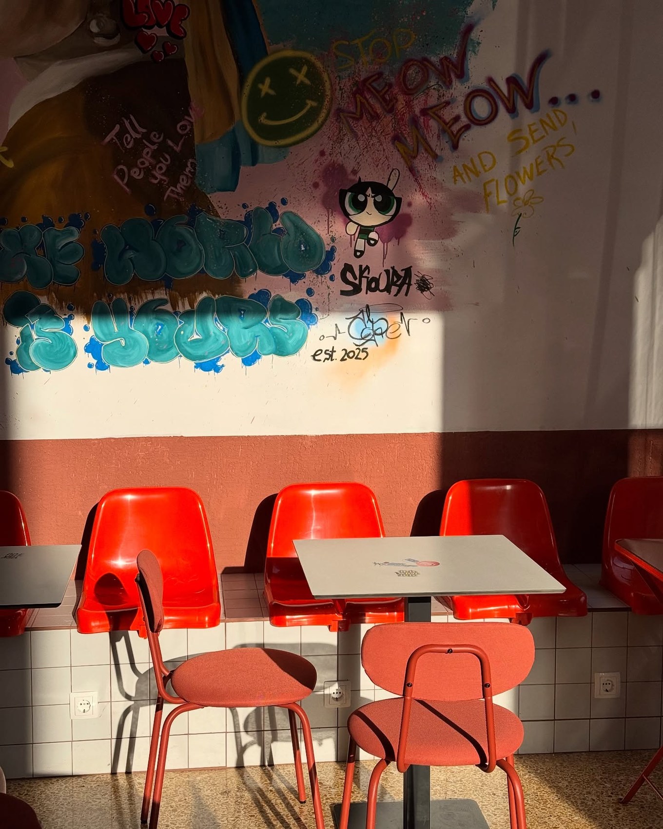 A colorful graffiti mural above orange and red chairs at an outdoor seating counter
