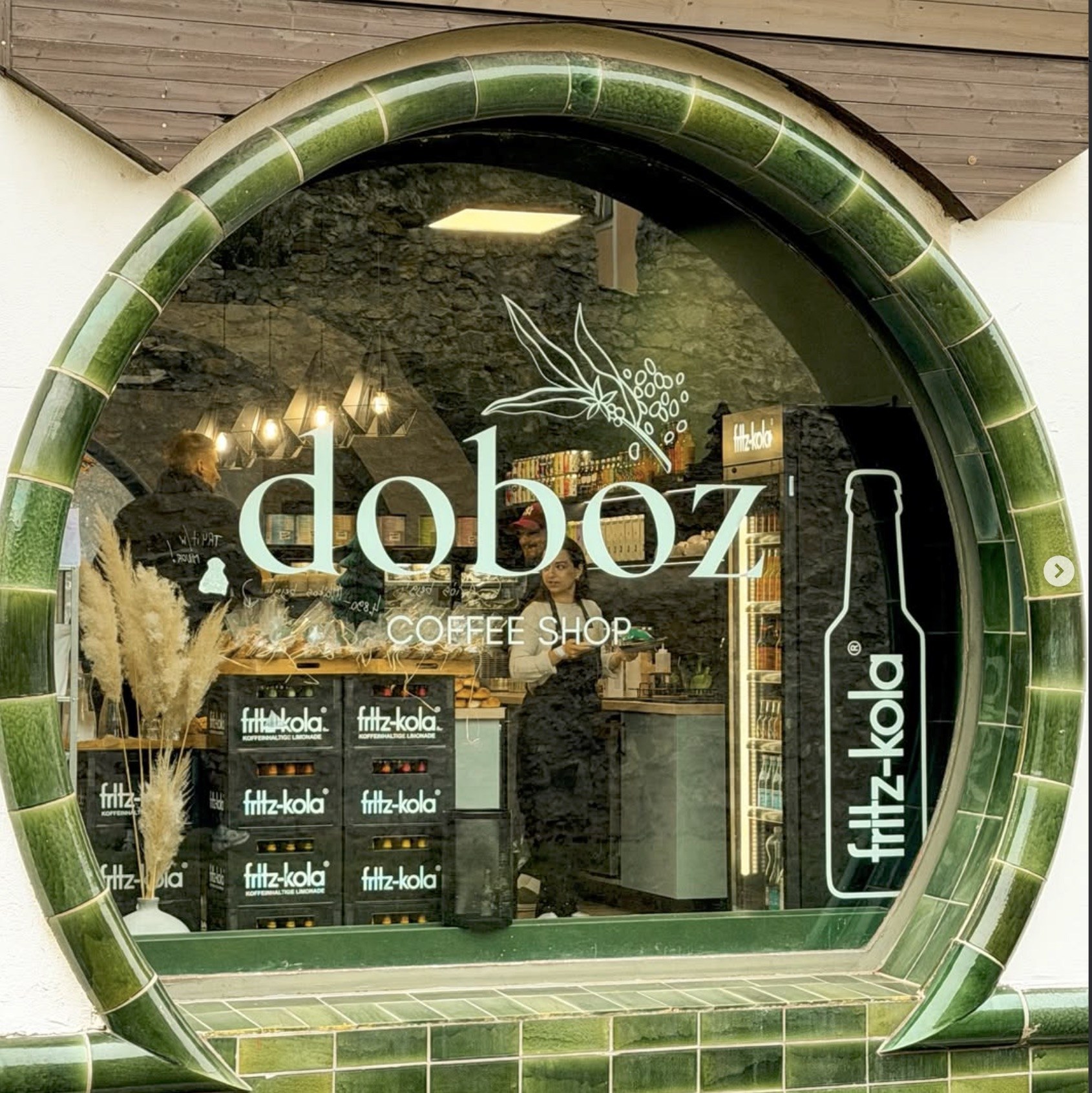 The round tiled window of Doboz Coffee Shop, with the shop's name etched into the glass and a staff member behind the counter inside