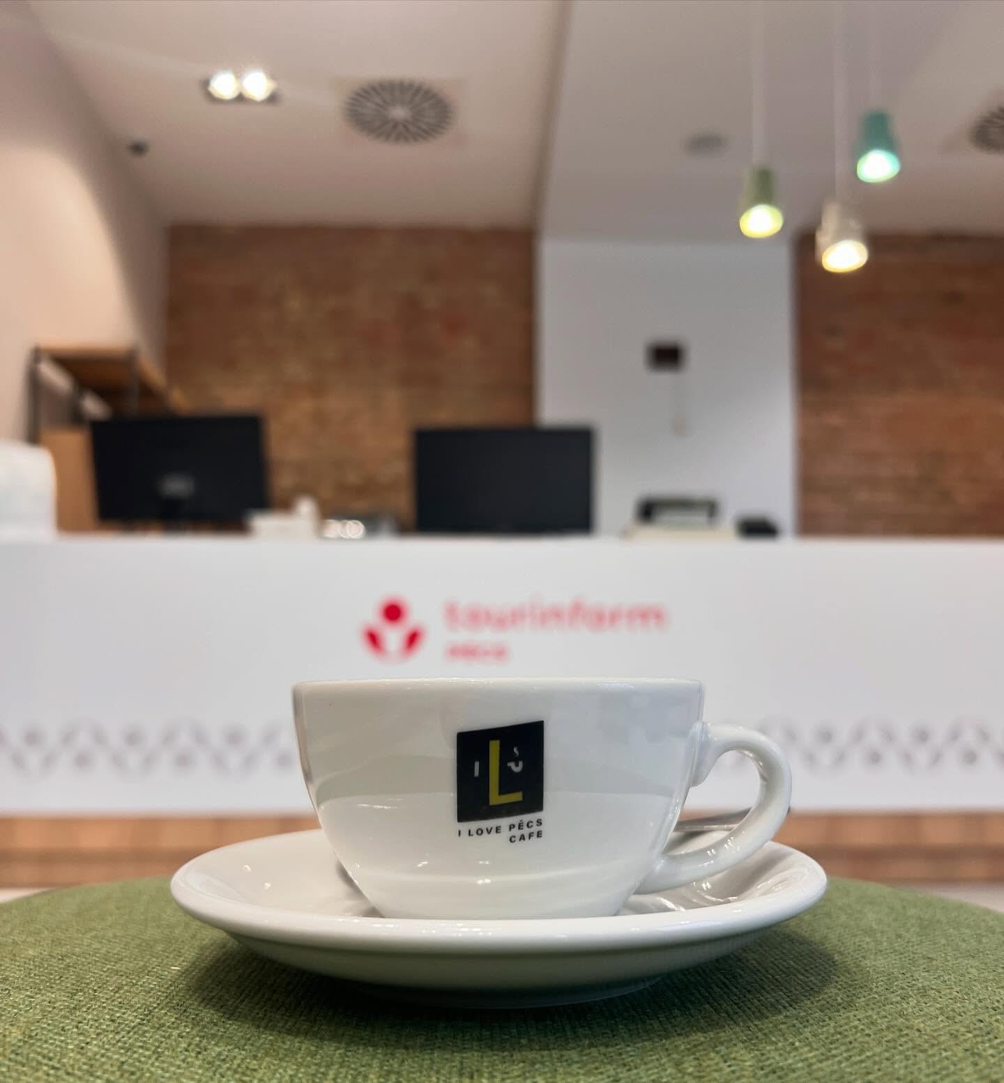 A branded I Love Pécs Café cup and saucer on a table, with the Pécs Point tourist information desk in the background