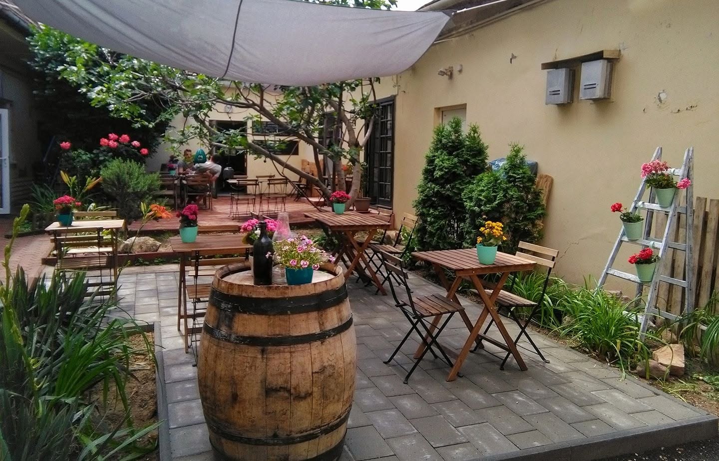 The shaded courtyard patio at Pécsi Kávé, with wooden tables, potted flowers, and a wine barrel used as a stand