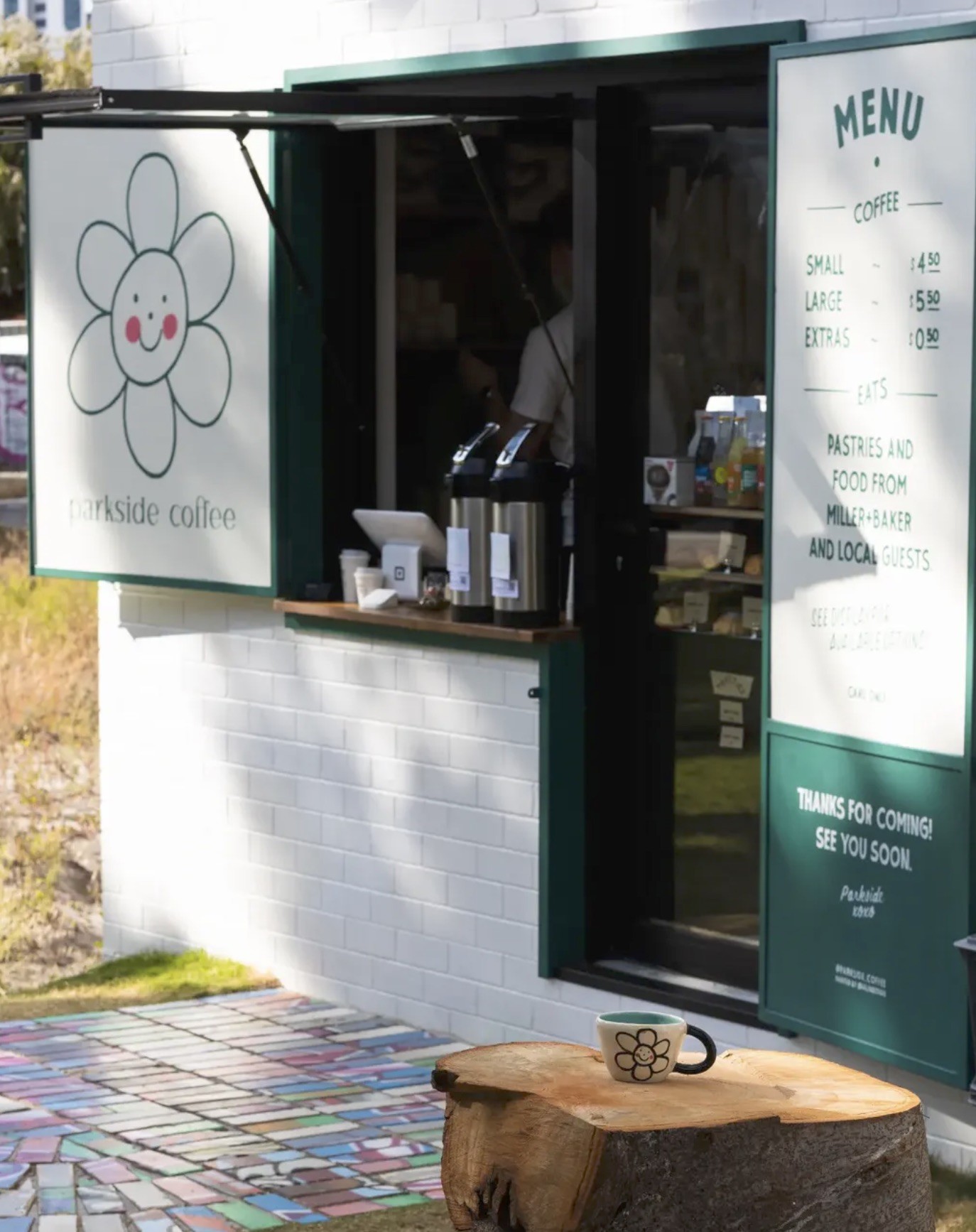 The Parkside Coffee hatch with its daisy sign and menu board, a branded mug on a tree stump table over the mosaic tiled ground out front