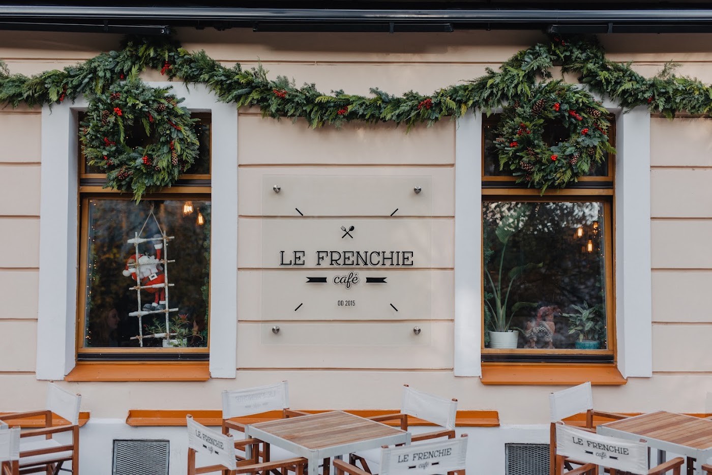 Le Frenchie cafe storefront with a wall-mounted sign reading Le Frenchie cafe between two garland-trimmed windows, and terrace tables set out front