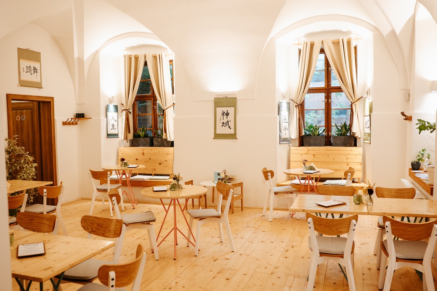 Bright vaulted dining room at Schovana Kavarna with pale wood tables and chairs, curtained windows, potted plants, and Japanese calligraphy scrolls on the walls