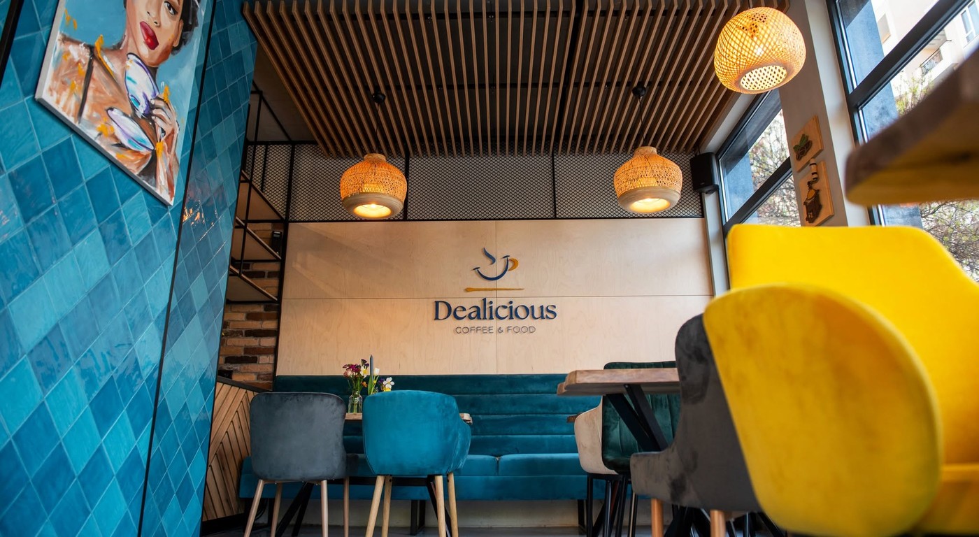 The Dealicious Coffee and Food logo mounted on a wood slat wall above teal velvet banquette seating, with pendant lamps and a colorful portrait painting on the tiled wall