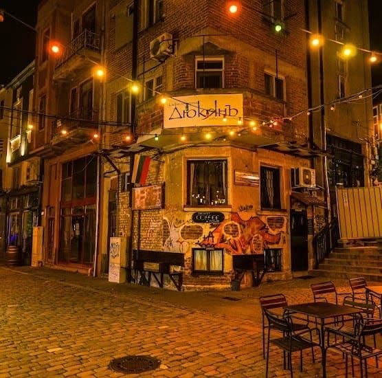 The corner storefront of Dyukyan Coffee Craftex at night, lit by string lights, with the shop's Cyrillic sign above a mural covered facade and empty outdoor tables on the cobblestones