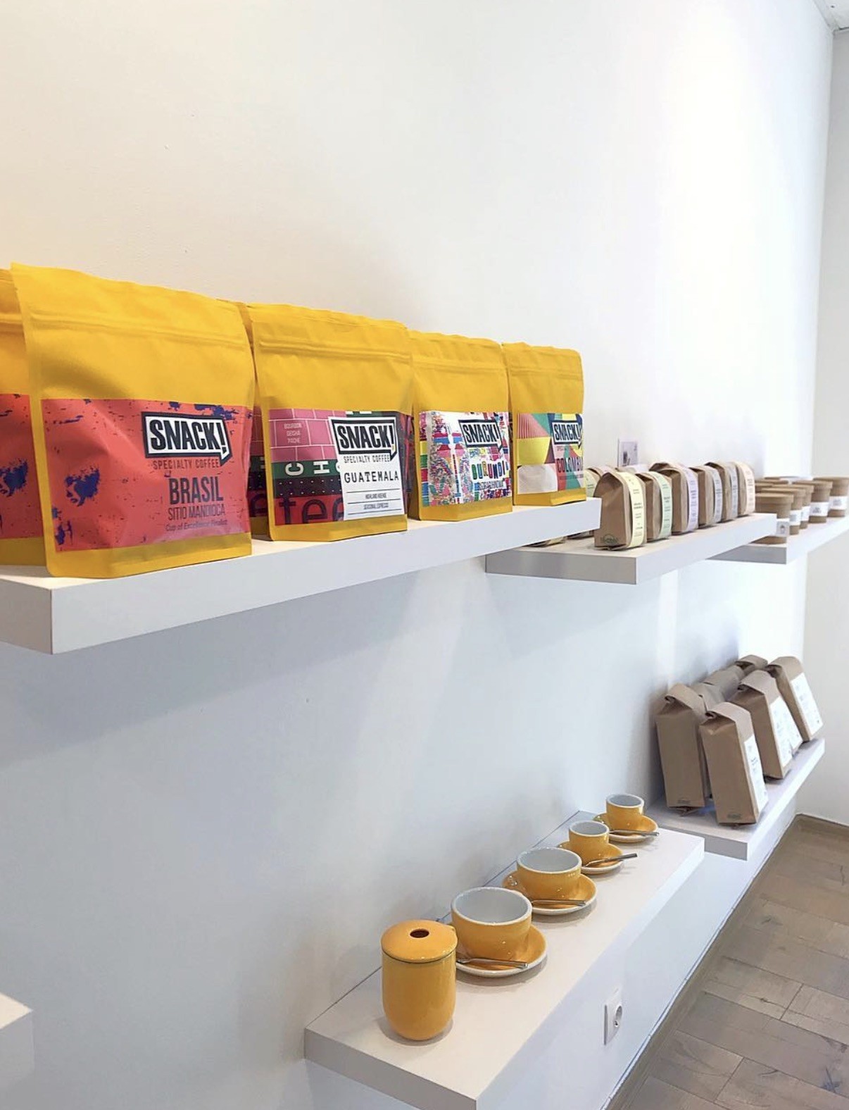 Yellow SNACK! Specialty Coffee bags lined up on a shelf above matching mustard yellow espresso cups and saucers