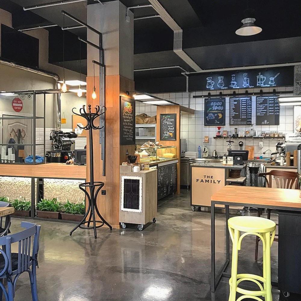 Interior view of The Family Coffee Roasters showing the wooden counter with THE FAMILY signage, an in-house roaster in the back corner, chalkboard menu boards and mixed seating