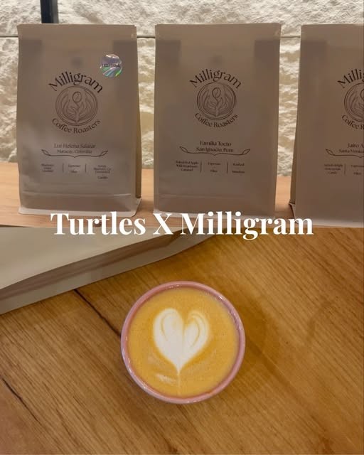 A latte with heart-shaped latte art in a pink cup on a wooden table, with bags of Milligram Coffee Roasters beans from a collaboration roast lined up behind it