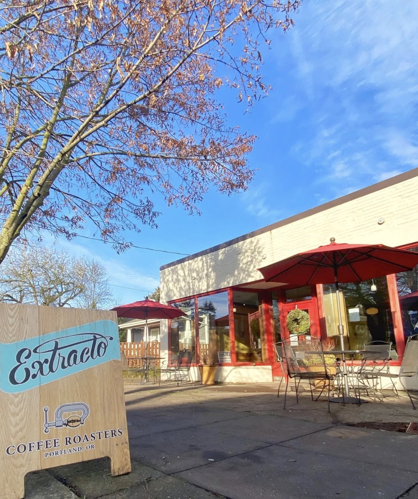 The wooden Extracto Coffee Roasters sidewalk sign in front of the red trimmed storefront with red patio umbrellas and outdoor seating