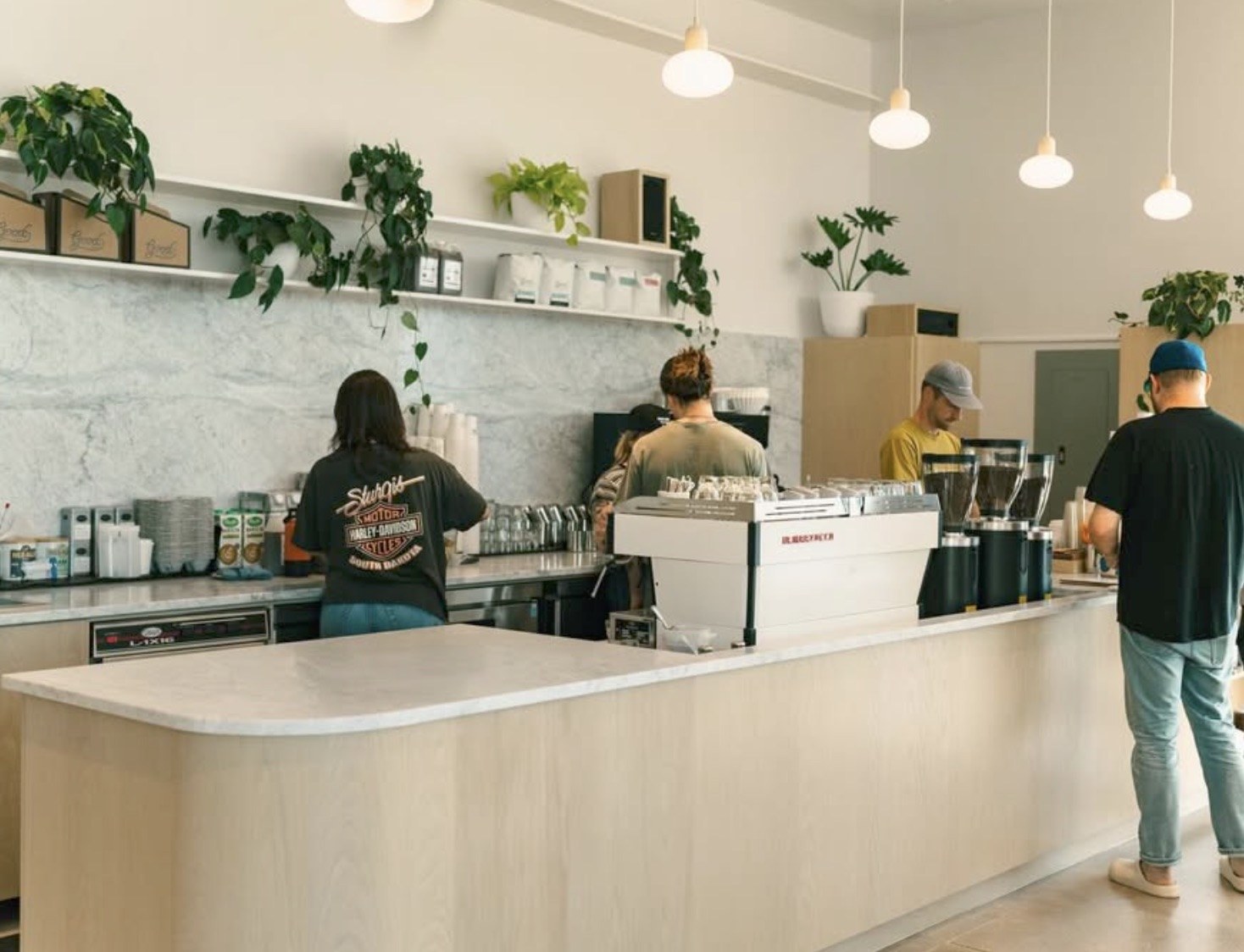 The marble espresso bar at Good Coffee, retail bean bags on a shelf above with plants and pendant lights overhead