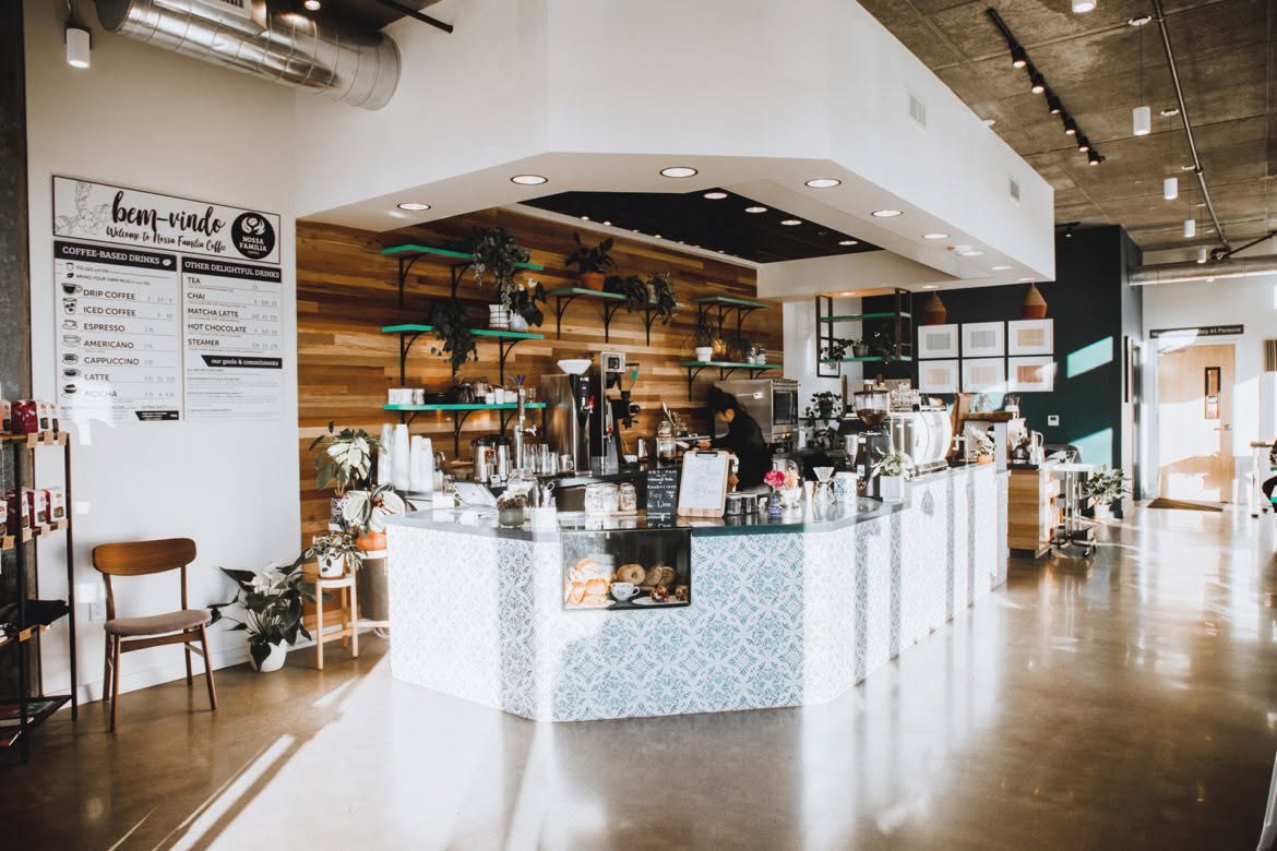 The Seven Corners cafe counter under a bem-vindo welcome sign, plants on green shelves and a barista working the espresso bar