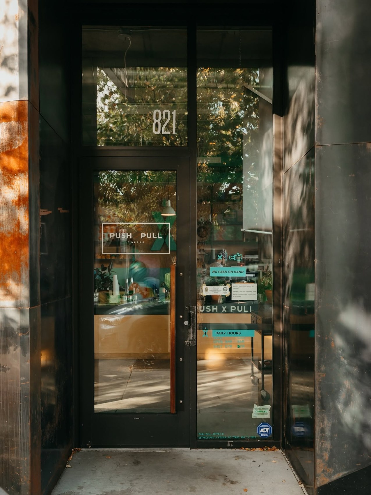 The glass front door at 821 with PUSH PULL COFFEE lettering and a teal X logo, daily hours posted beside it