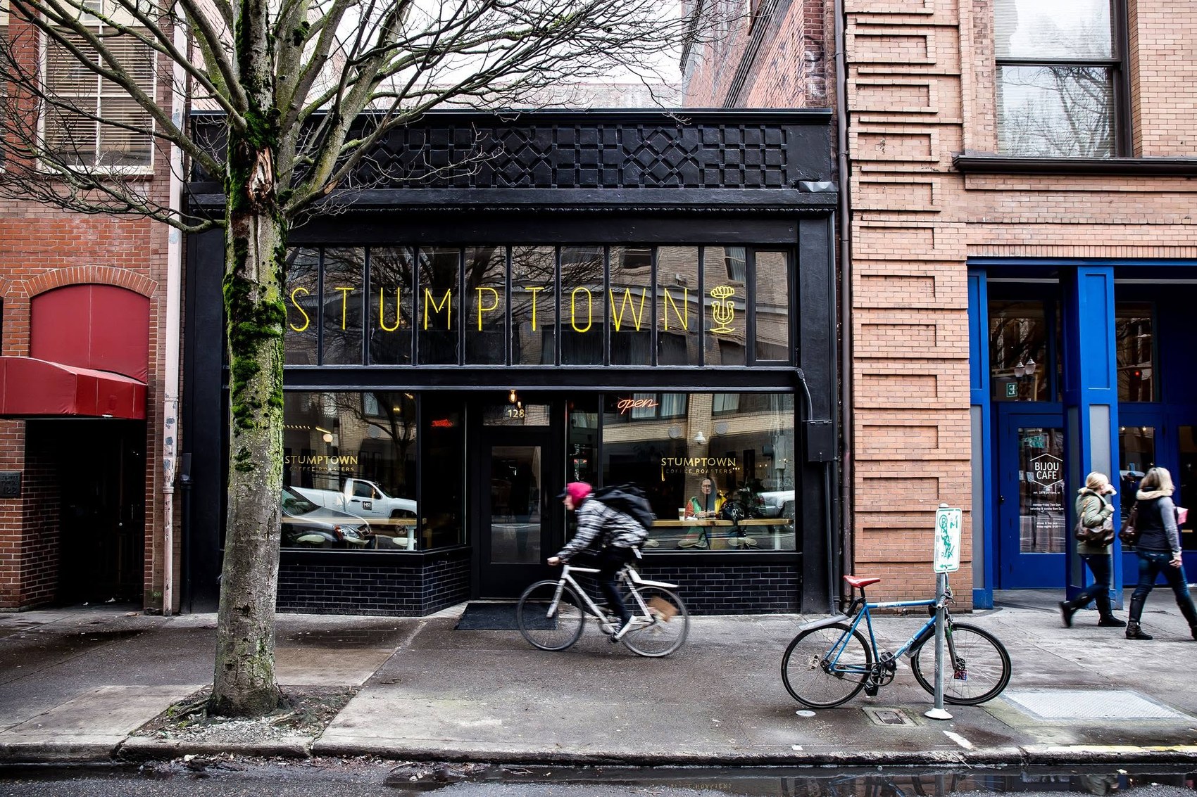 A black storefront with a yellow STUMPTOWN sign in the window, a cyclist passing on the sidewalk out front