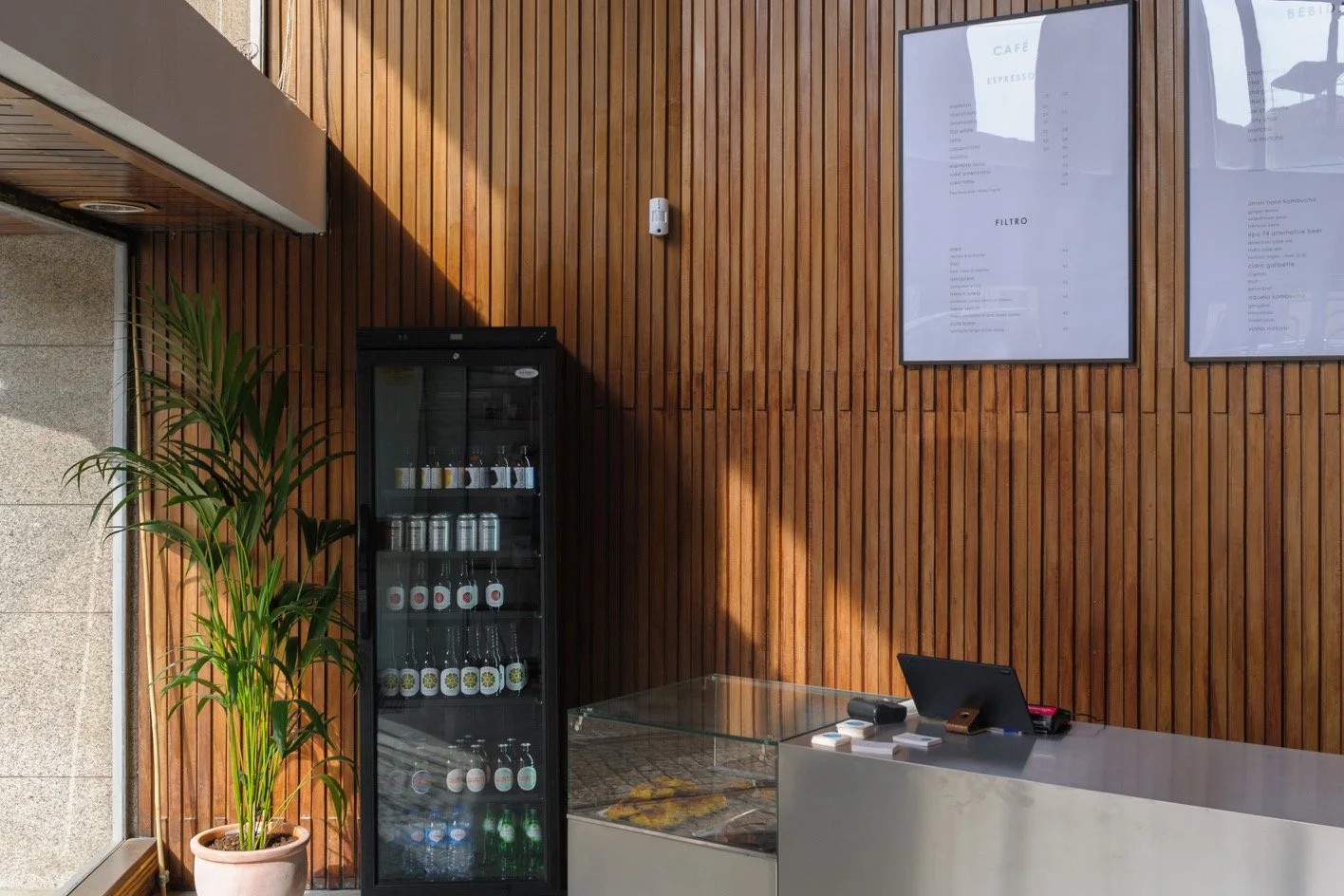 The wooden slat wall at SO Coffee Roasters on Rua da Restauração, framed espresso and filter menus above a stainless steel counter with a pastry case and a drinks fridge beside a potted palm