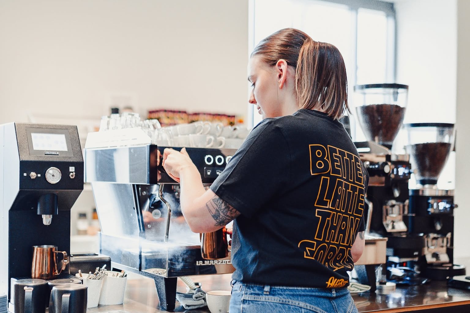 A barista steaming milk on a La Marzocco espresso machine between two large coffee grinders at EMA espresso bar
