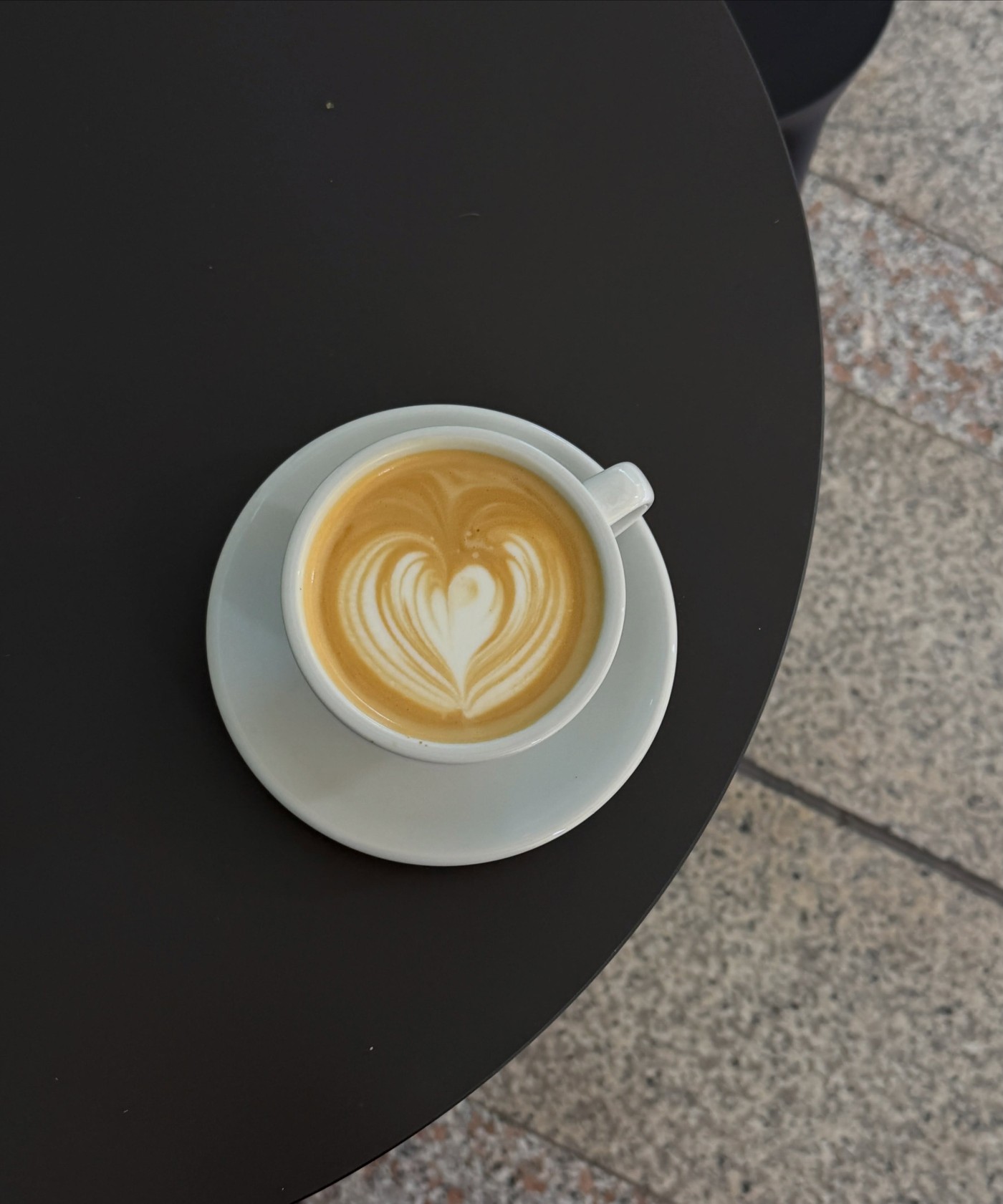 A flat white with heart shaped latte art on a white cup and saucer, sitting on a round black table with a terrazzo floor in the background