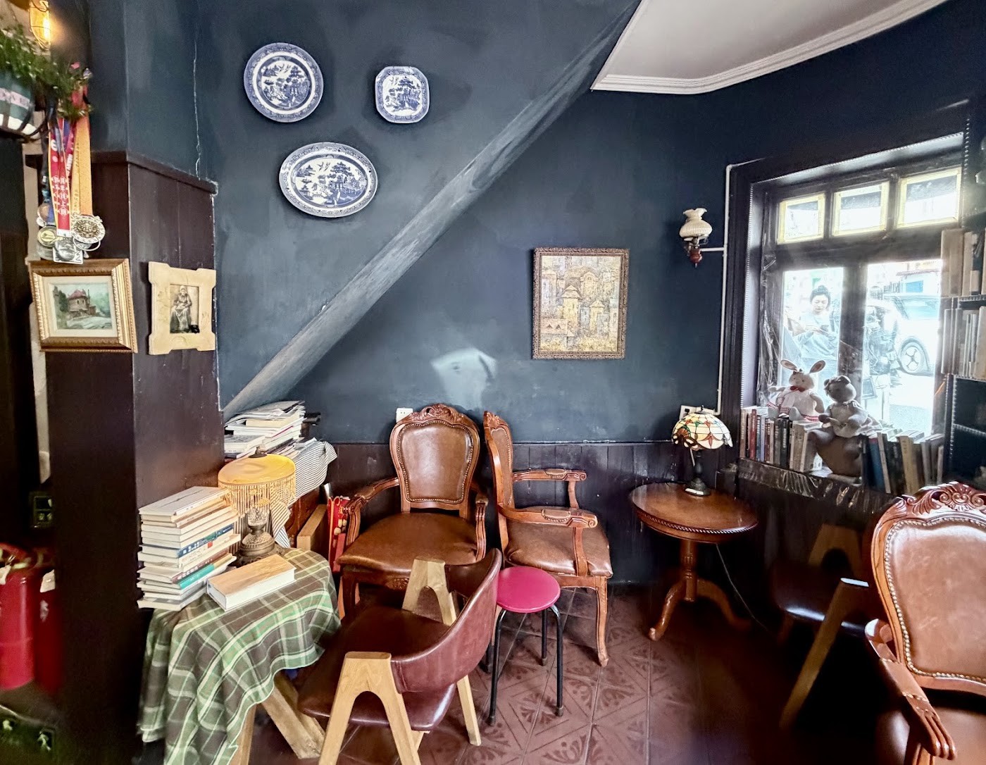 A corner of the dark blue walled interior with antique leather armchairs, a round wood table, stacked books, a stained glass lamp and blue and white porcelain plates on the wall