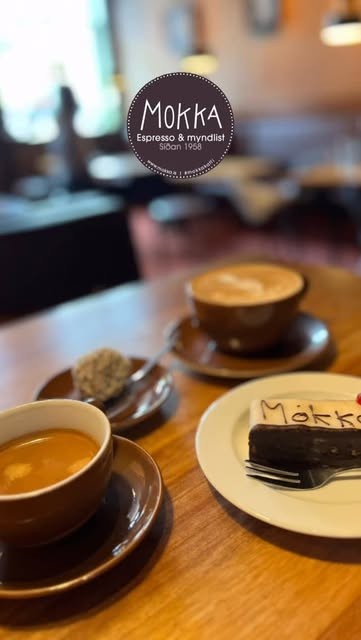 Two espressos, a cappuccino, and a branded slice of Mokka chocolate cake on a wooden table beneath the cafe's logo
