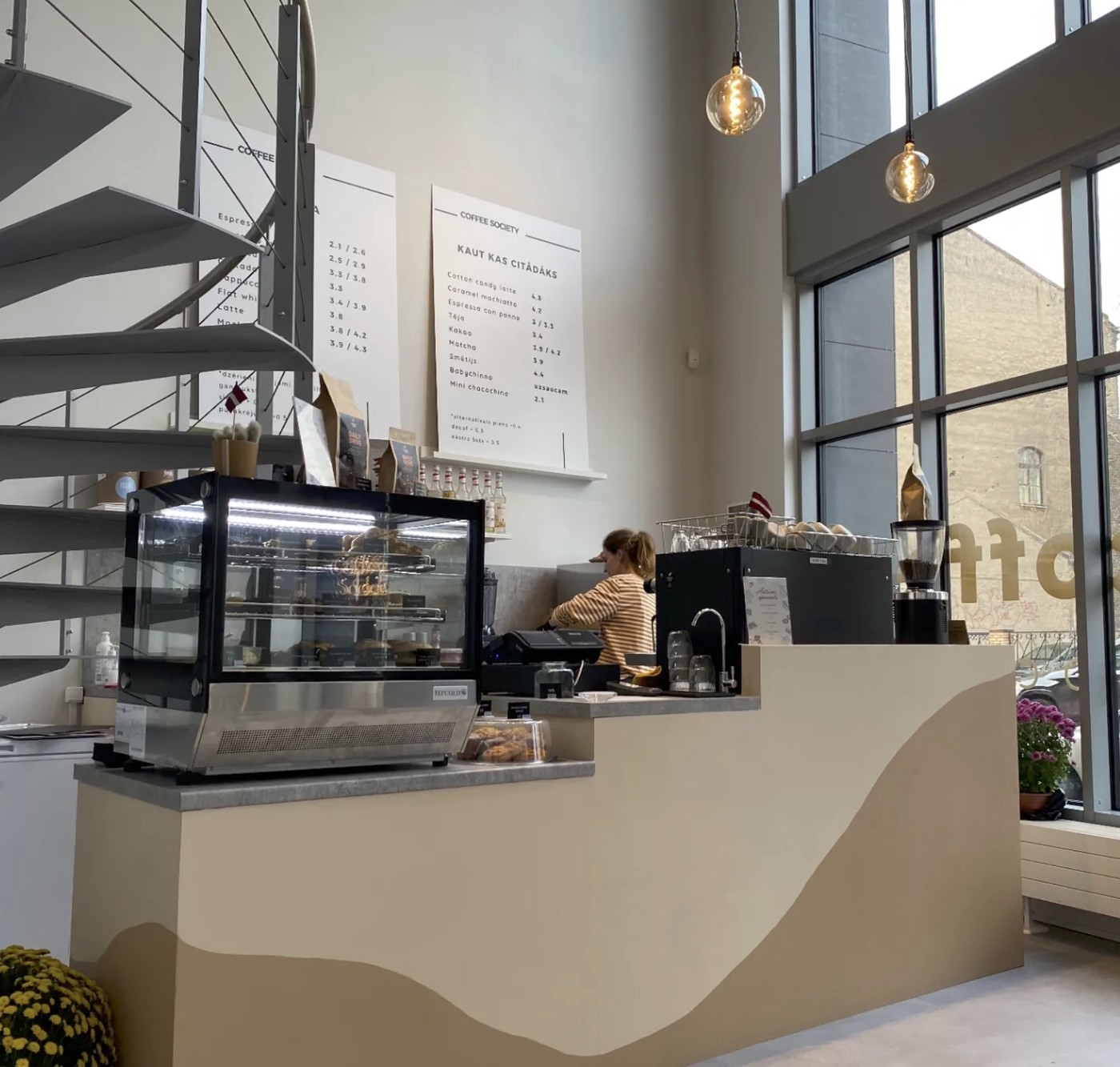 The Coffee Society counter and pastry case beneath a hand lettered menu board, a barista working with her back to the camera