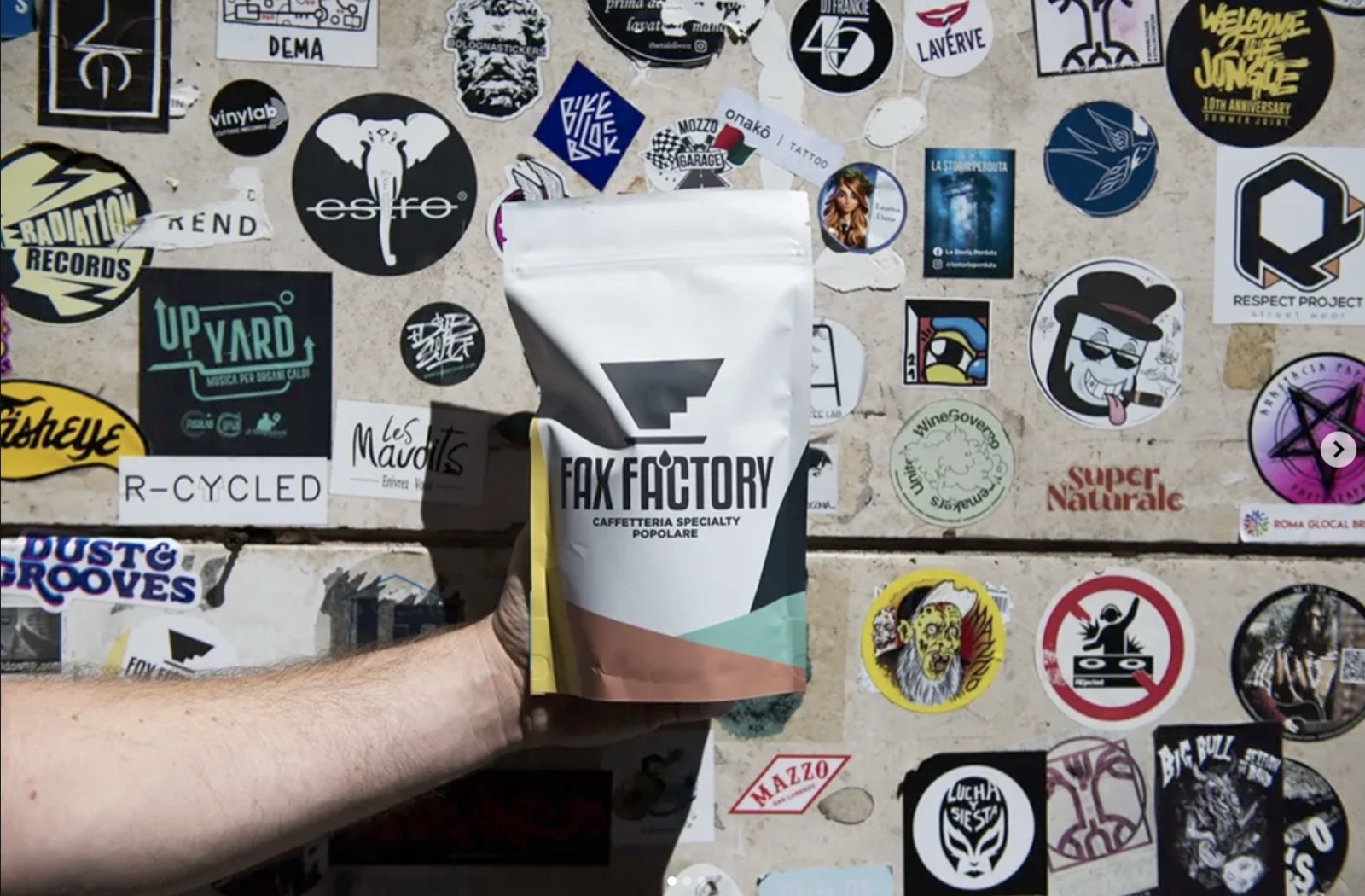 A branded Fax Factory 'Caffetteria Specialty Popolare' coffee bag held up against a wall covered in stickers