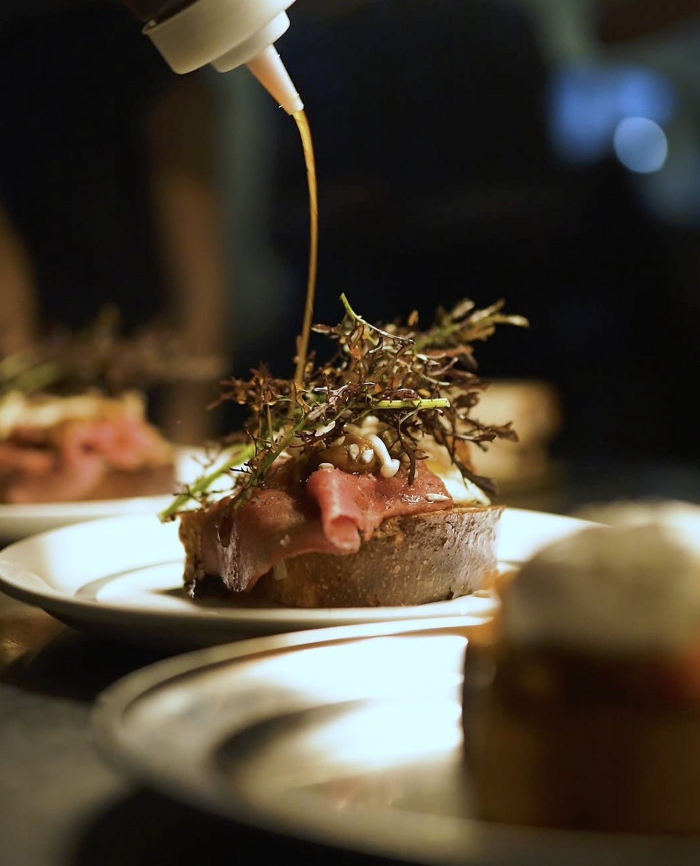 Oil being drizzled over an open faced toast topped with prosciutto and microgreens on a plate at Harvest Cafe & Bakery