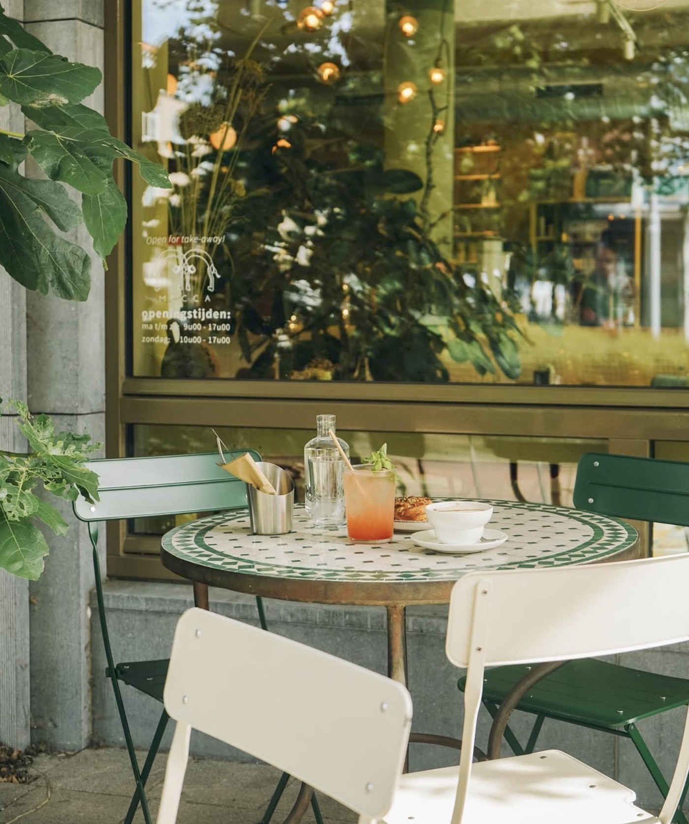 An outdoor mosaic tile table set with a coffee, a pink drink, and a pastry outside Oasis's storefront window