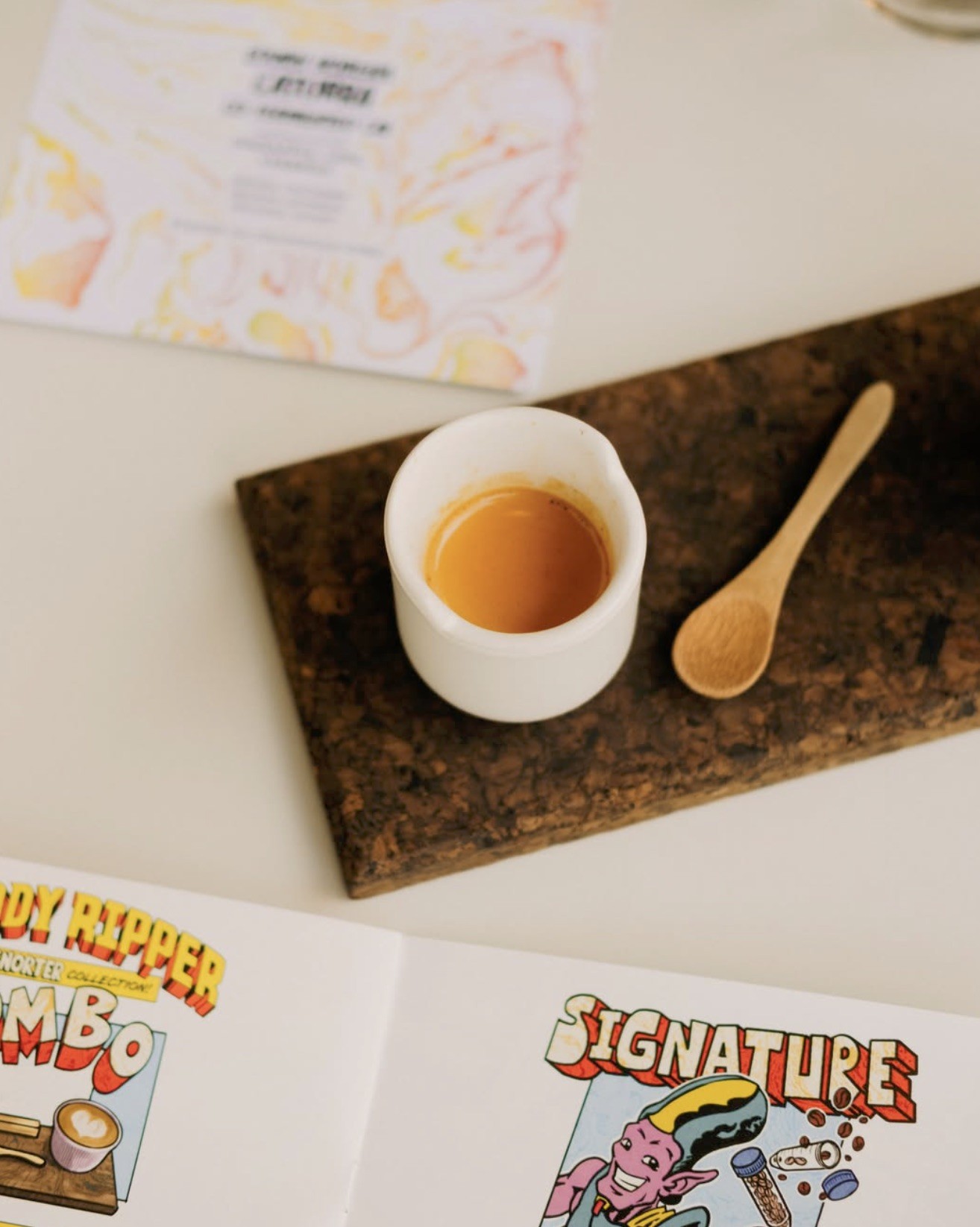 An espresso in a small white cup with a wooden spoon on a cork board beside the Bloody Ripper Collection menu at Ripsnorter