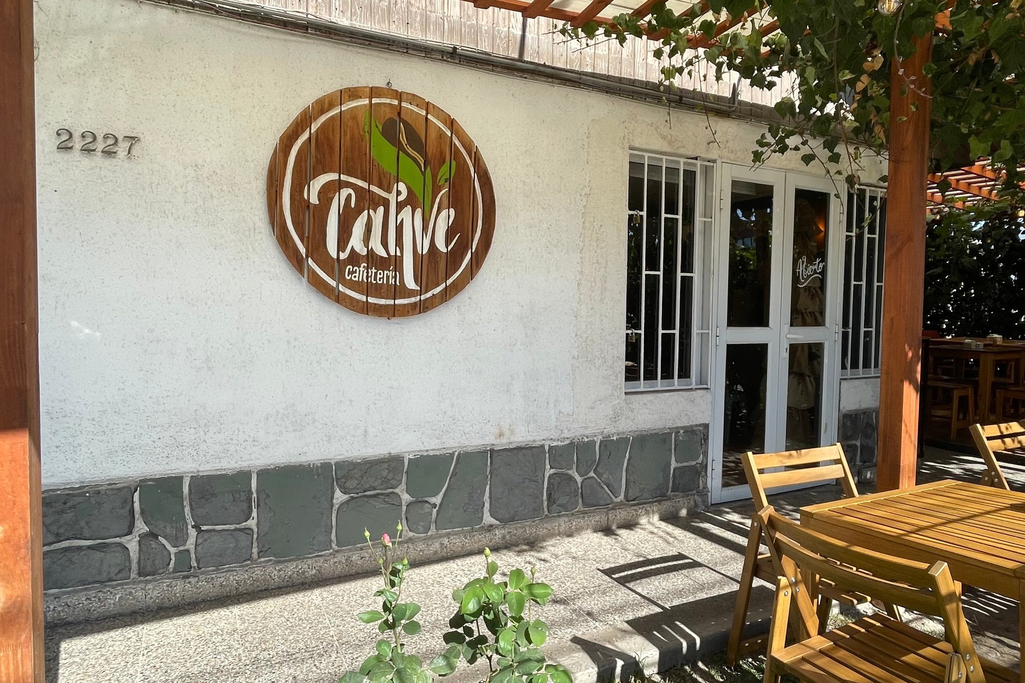 The whitewashed storefront of Cahve Cafetería in Maipú with its round wooden sign, street number 2227 and wooden tables under a pergola terrace