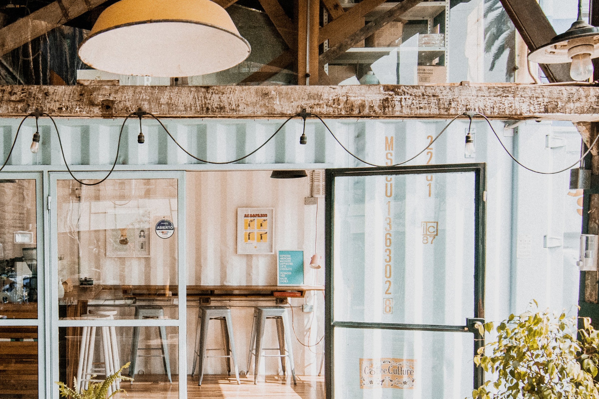The pale blue shipping container cafe of Coffee Culture Coffee Roasters at Av. La Paz with string lights, a high wooden bar and metal stools visible through the windows
