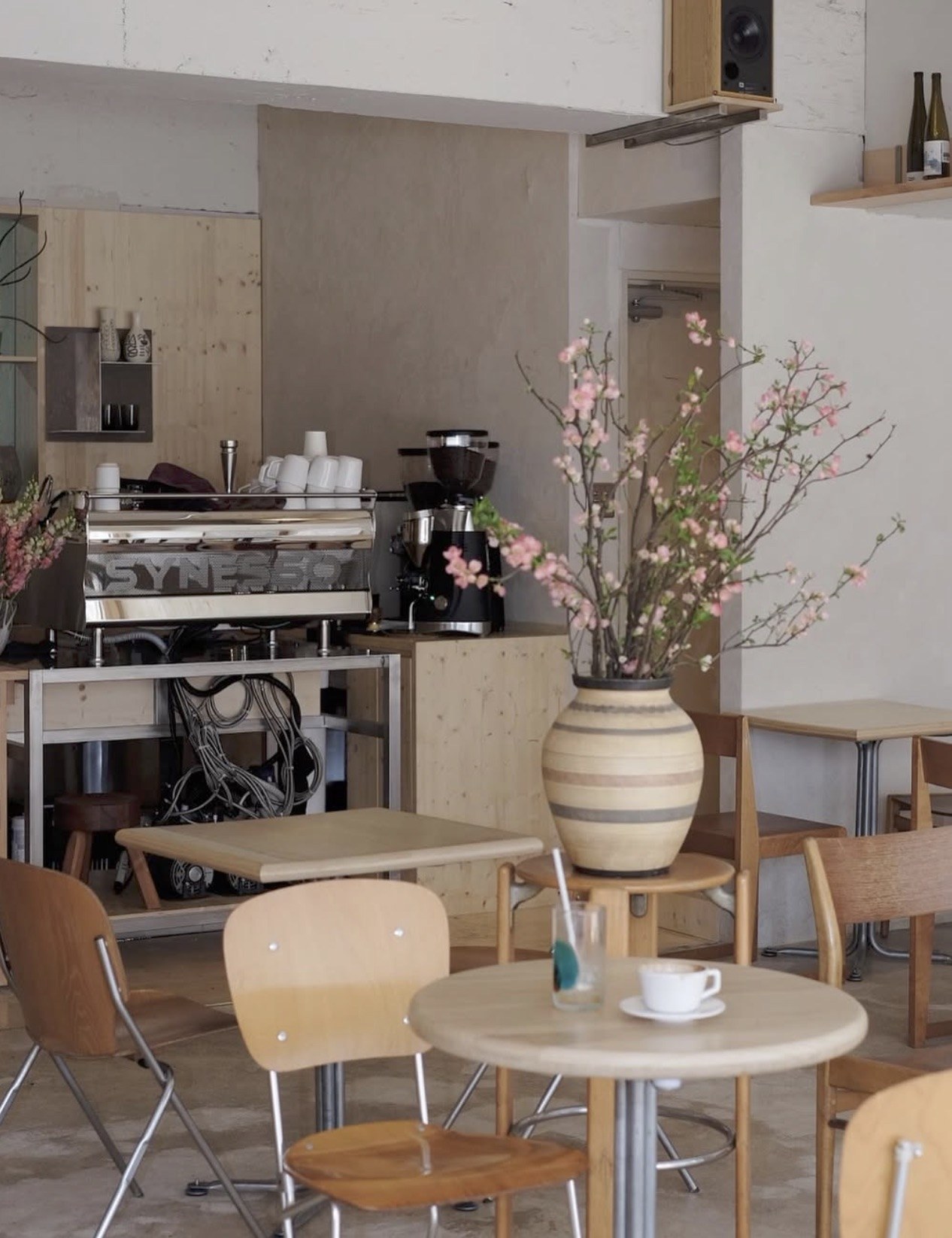 The espresso bar at Always August with a Synesso machine and grinder, round wood tables and a large vase of cherry blossom branches in the foreground