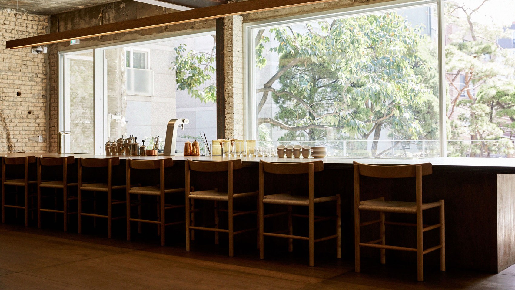 A long wooden bar with woven-seat stools lined up under floor to ceiling windows looking out on trees, exposed brick to one side