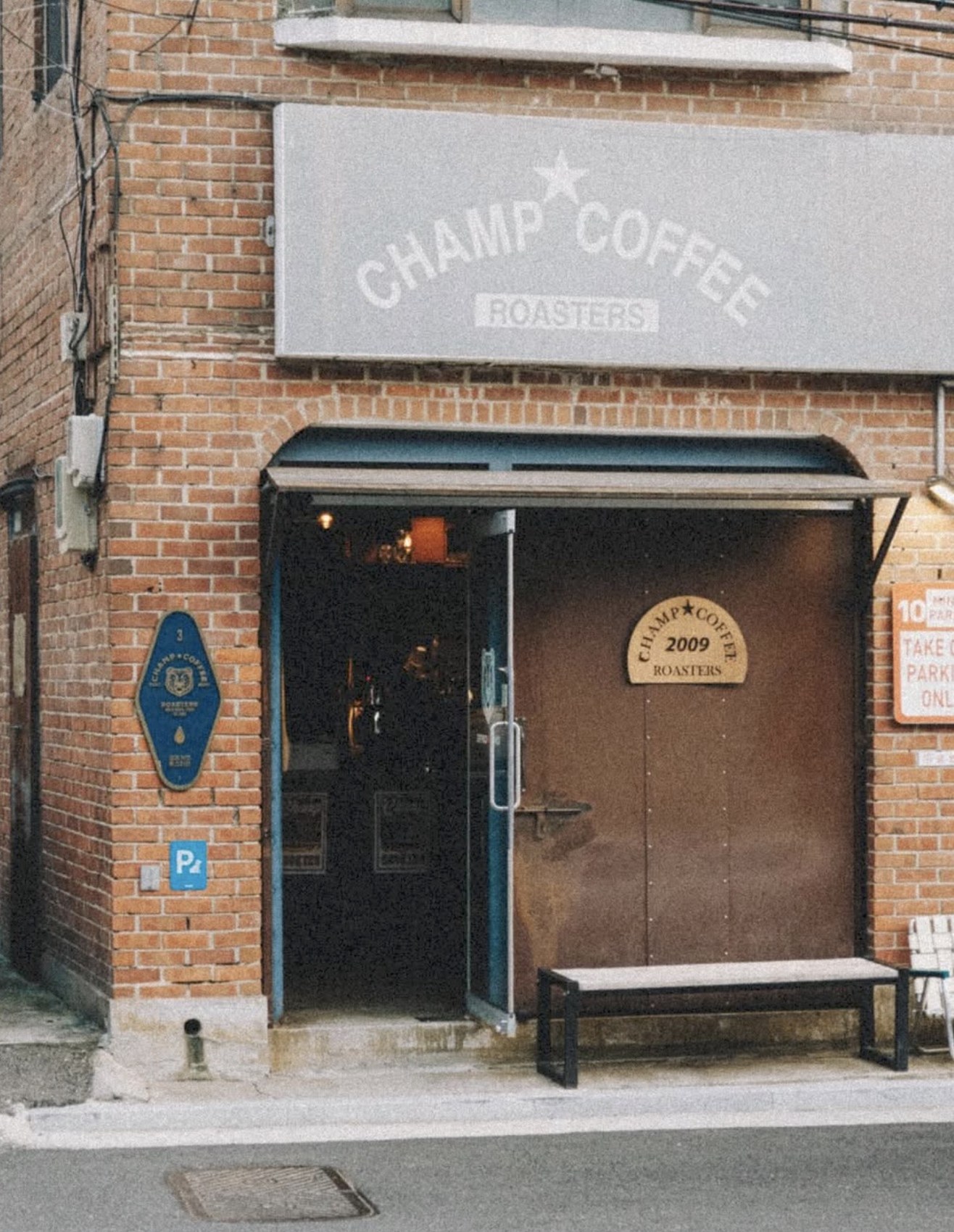 The brick storefront of Champ Coffee Roasters, a CHAMP COFFEE sign above an open door and a 2009 founding plaque beside a bench